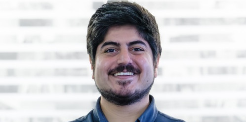 Eine Person steht vor einem unscharfen Hintergrund mit horizontalen hellen und dunklen Streifen. Die Person hat dunkles, kurzes Haar, einen Vollbart und einen Schnurrbart. Sie trägt ein dunkelblaues Hemd mit Stehkragen und Knopfleiste. Die Person lächelt freundlich und schaut direkt in die Kamera.