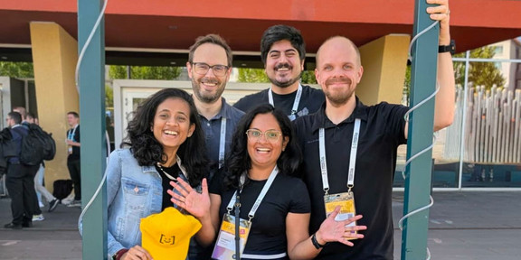 The image has five people standing behind a frame assigned for taking pictures with the conference name to tag it on social media