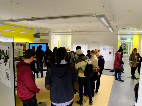 Approximately 20 people in a small atrium area, holding conversations, viewing posters, and trying out demos. 