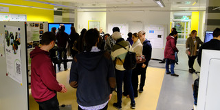 Approximately 20 people in a small atrium area, holding conversations, viewing posters, and trying out demos. 