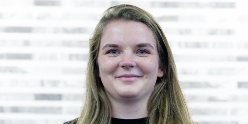 Eine Person steht vor einem unscharfen Hintergrund mit horizontalen hellen und dunklen Streifen. Die Person hat dunkles, kurzes Haar, einen Vollbart und einen Schnurrbart. Sie trägt ein dunkelblaues Hemd mit Stehkragen und Knopfleiste. Die Person lächelt freundlich und schaut direkt in die Kamera.