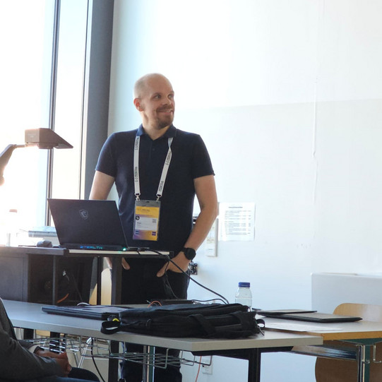 The image shows a person smiling, standing behind a podium with a laptop giving a presentation