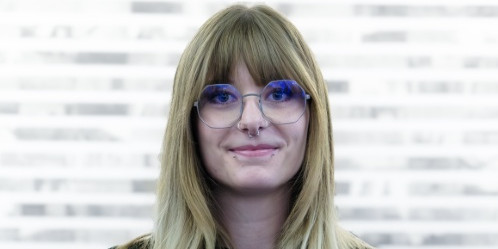 Eine Person steht vor einem unscharfen Hintergrund mit horizontalen hellen und dunklen Streifen. Die Person hat dunkles, kurzes Haar, einen Vollbart und einen Schnurrbart. Sie trägt ein dunkelblaues Hemd mit Stehkragen und Knopfleiste. Die Person lächelt freundlich und schaut direkt in die Kamera.
