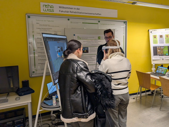 One person wears a VR headset to try out a simulation with the help of one of the students from our project. Two other onlookers watch the interaction on a large screen nearby. 