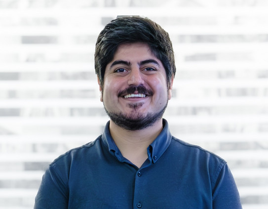 Eine Person steht vor einem unscharfen Hintergrund mit horizontalen hellen und dunklen Streifen. Die Person hat dunkles, kurzes Haar, einen Vollbart und einen Schnurrbart. Sie trägt ein dunkelblaues Hemd mit Stehkragen und Knopfleiste. Die Person lächelt freundlich und schaut direkt in die Kamera.